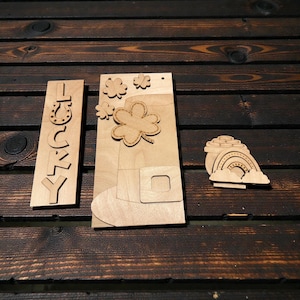 May include: Wooden St. Patrick's Day decorations. Includes a vertical sign with the word "Lucky", a leprechaun hat with shamrocks, and a pot of gold with a rainbow. The items are made of light-colored wood and are displayed on a dark wood surface.