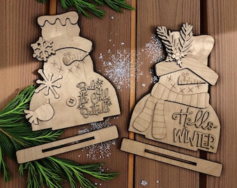 May include: Two wooden snowman-shaped decorations. One has a snowflake, bird, and text that reads "Baby it's Cold Outside." The other has a scarf, hat, and text that reads "Hello Winter." Both have a stand.