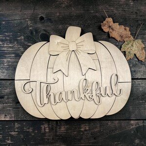 May include: A light-colored wooden pumpkin cutout featuring a decorative bow at the top and the word Thankful written in a cursive script across the center. The pumpkin is constructed from multiple horizontal wood panels with visible seams.