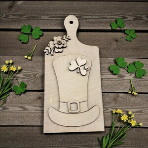 May include: A wooden cutting board with a handle, decorated with a leprechaun hat and shamrock design. The board is surrounded by green shamrocks and yellow flowers, perfect for St. Patrick's Day decor.