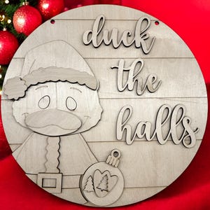 May include: A round wooden Christmas sign with a Santa Claus duck design and the words "duck the halls". The sign is made of light-colored wood and features a decorative ornament with trees.