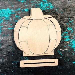 May include: A light-colored wooden pumpkin cutout featuring a segmented design with visible curved lines. The pumpkin is attached to a flat, rectangular base with a horizontal slot, allowing the object to stand upright on a flat surface.