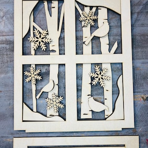 May include: A wooden cutout of a window with four panes, each depicting a winter scene with birch trees, snowflakes, and birds. The cutout is designed to be used as a decorative element for a holiday project.