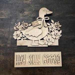 May include: Wooden laser-cut goose with floral accents and a tag that says "Honk Honk." The goose is surrounded by flowers with button centers. Below is a separate wooden sign that reads "Hey Silly Goose."