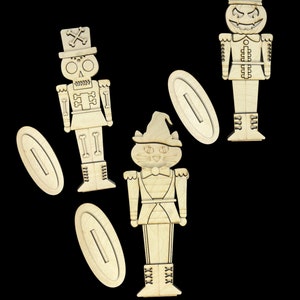 May include: Three wooden nutcracker ornaments, one with a skull, one with a pumpkin face, and one with a cat face. Each ornament has a slot for a small gift card or note.