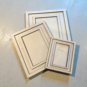 May include: Four unfinished wooden picture frames of varying sizes are arranged on a light gray surface. The frames have a natural wood color with a dark border, creating a layered effect. The frames are rectangular and ready for customization.