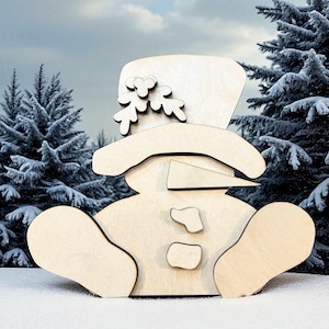 May include: A wooden snowman figure with a top hat and twig accents. The snowman is sitting in the snow with a wintery background of snow-covered evergreen trees and a cloudy sky. The snowman's nose is a triangle.