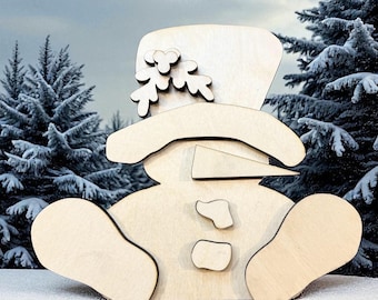 Puede incluir: Una figura de muñeco de nieve de madera con sombrero de copa y adornos de ramitas. El muñeco de nieve está sentado en la nieve con un fondo invernal de árboles de hoja perenne cubiertos de nieve y un cielo nublado. La nariz del muñeco de nieve es un triángulo.