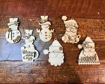 May include: A collection of unfinished wooden Christmas ornaments. Designs include snowmen, Santa figures, and a mug with the words "Warm and Cozy." Each ornament has a small hole for hanging.
