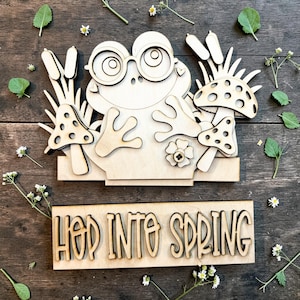 May include: Wooden frog-themed spring decoration with the text "HOP INTO SPRING". The frog wears glasses and is surrounded by mushrooms and cattails. The decoration is made of light-colored wood and is set against a wooden background.