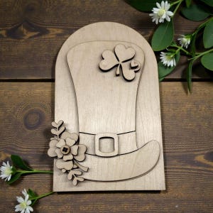 May include: Wooden St. Patrick's Day decoration featuring a leprechaun hat with a shamrock and floral accents. The hat has a buckle on the band. The decoration is made of light-colored wood and is set against a dark wood background.