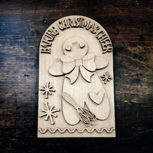 May include: A light-colored wooden arch-shaped sign featuring a gingerbread person with a bow tie, a heart, and a whisk. The text BAKING CHRISTMAS CHEER is engraved at the top, accompanied by star-shaped accents and a wavy line at the bottom edge.
