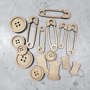 May include: A collection of wooden craft supplies, including buttons, safety pins, and spools. The buttons have three or four holes. The safety pins are in various sizes. The spools are rectangular with curved edges. All items are made of light-colored wood.