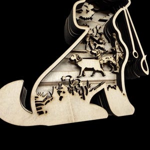 May include: A layered wooden art piece depicting a hunting scene. The artwork features a silhouette of a dog with a hunter and dogs in a forest setting. The piece is made of light-colored wood with a dark background.