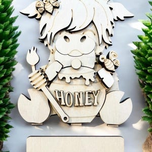 May include: Wooden honey-themed decoration featuring a cow with a honey pot and a honey dipper. The word "HONEY" is carved into the pot. Includes a separate wooden base with three slots. Decorative bees and floral accents are also present.