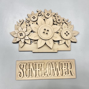 May include: A light-colored wooden cutout featuring a cluster of sunflower shapes with four-hole buttons at their centers, attached to a rectangular base. Below this, a separate rectangular wooden sign displays the word SUNFLOWER in cutout letters.