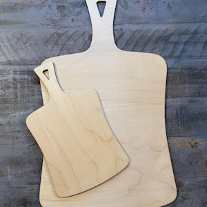 May include: Two wooden cutting boards, one smaller than the other, with handles. The larger board is light brown and the smaller board is a slightly darker shade of brown.