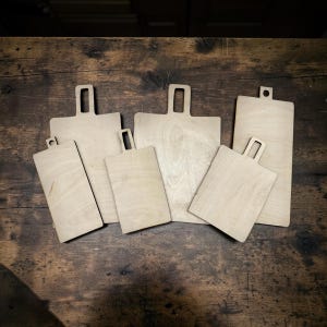 May include: A collection of five light-colored wooden cutting boards of varying rectangular sizes. Each board features a flat, smooth surface and a handle with a cutout hole at the top, arranged in a cluster on a dark, rustic wooden surface.