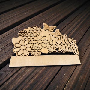 May include: Wooden sign with the words "hello SPRING" cut out, along with flower and butterfly designs. The sign has a rectangular base for a name or message. The sign is made of light-colored wood and sits on a dark wood surface.