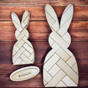May include: Two wooden bunny shapes with a woven pattern. The larger bunny is standing upright, while the smaller bunny is lying down. The bunnies are on a wooden surface.