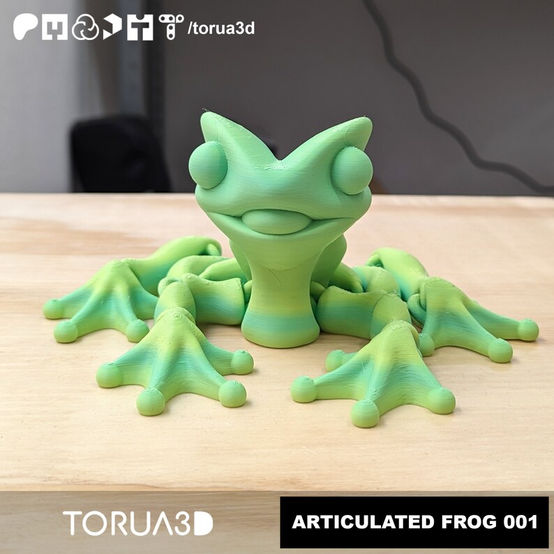 May include: A green articulated frog figurine with a textured surface. The frog has large eyes and a wide smile. The figurine is posed sitting on a wooden surface. The text "ARTICULATED FROG 001" is printed on the surface below the frog.