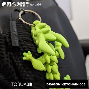 May include: A bright green dragon keychain with a silver keyring attached to a black bag. The keychain is 3D-printed and has the text "DRAGON KEYCHAIN 005" in a white box. The brand name "TORUA3D" is also visible.
