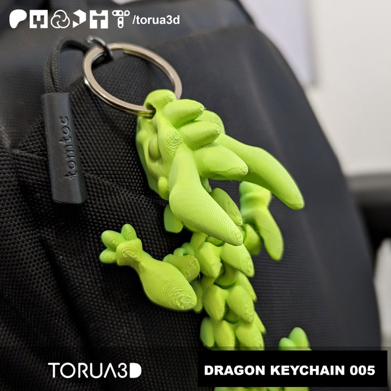 May include: A bright green dragon keychain with a silver keyring attached to a black bag. The keychain is 3D-printed and has the text "DRAGON KEYCHAIN 005" in a white box. The brand name "TORUA3D" is also visible.