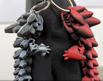 May include: Two articulated dragon keychains, one gray and one red, attached to a silver key ring. The dragons are made of 3D printed plastic and have a textured surface.