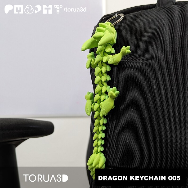 May include: A lime green dragon keychain made of articulated segments hangs from a silver keyring attached to a black backpack. The keychain is labeled "DRAGON KEYCHAIN 005" and the brand is "TORUA3D".