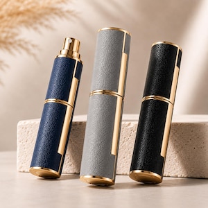 Könnte beinhalten: Drei zylindrische Parfümzerstäuber mit strukturierten Kunstlederhüllen in Marineblau, Grau und Schwarz. Jeder Zerstäuber verfügt über einen polierten Goldmetallrand, einen passenden goldenen Boden und eine goldene Sprühdüse am blauen Flakon.