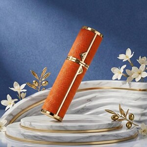 May include: An orange, textured perfume atomizer with gold accents. The cylindrical container has a gold band and geometric design. The atomizer is displayed on a marble platform with white flowers and gold decorative elements.