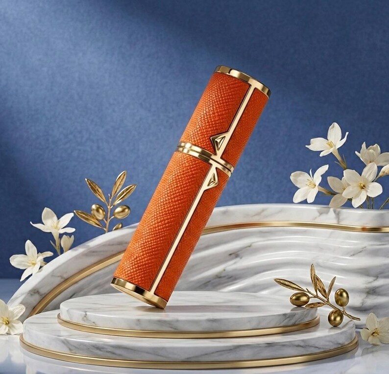 May include: An orange, textured perfume atomizer with gold accents. The cylindrical container has a gold band and geometric design. The atomizer is displayed on a marble platform with white flowers and gold decorative elements.