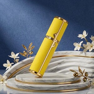 May include: A yellow, rectangular perfume atomizer with gold accents. The atomizer has a textured surface and is decorated with gold trim and a gold clasp. The product is displayed on a marble platform with floral and olive branch decorations.