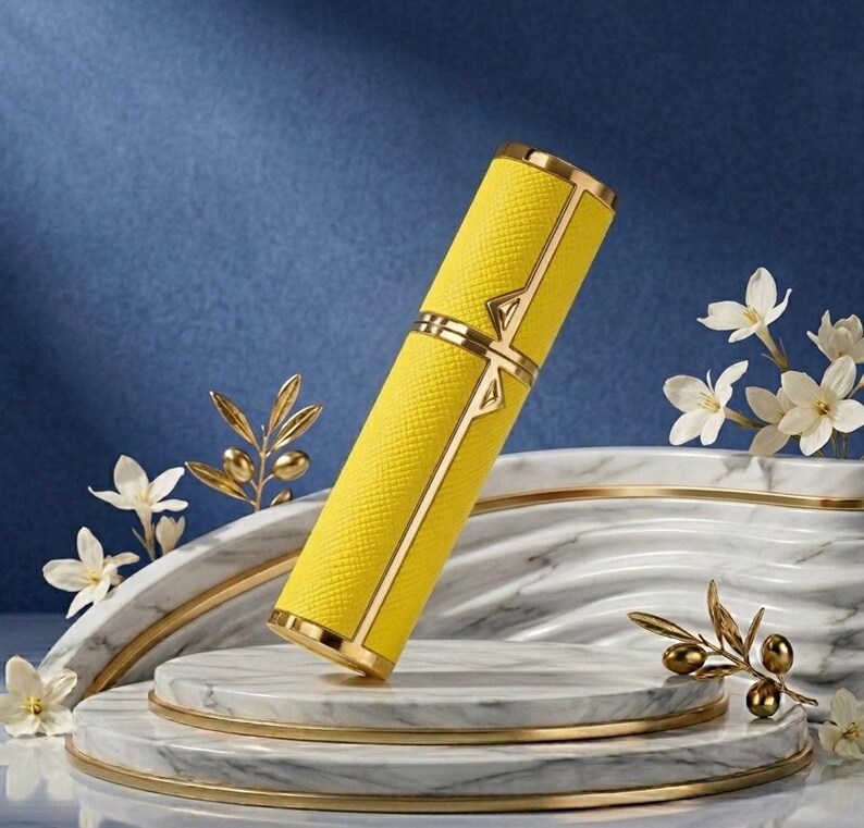 May include: A yellow, rectangular perfume atomizer with gold accents. The atomizer has a textured surface and is decorated with gold trim and a gold clasp. The product is displayed on a marble platform with floral and olive branch decorations.