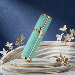 May include: A teal-colored, rectangular perfume bottle with gold accents. The bottle has a textured surface and is decorated with gold geometric designs. The bottle is displayed on a marble platform with gold trim, with white flowers and gold foliage in the background.
