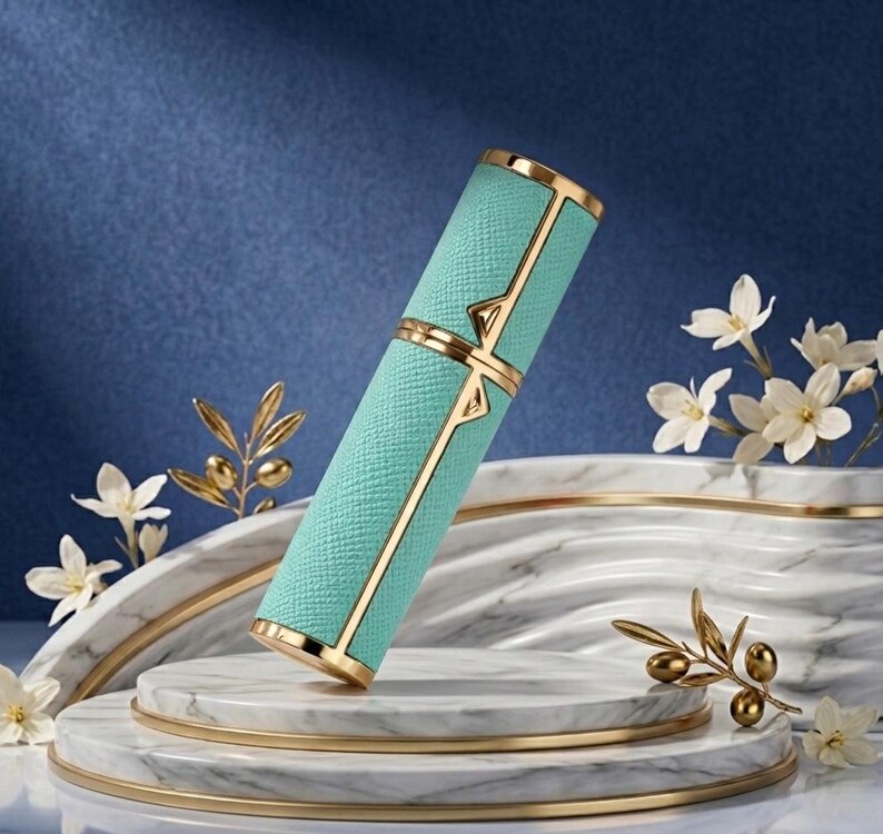 May include: A teal-colored, rectangular perfume bottle with gold accents. The bottle has a textured surface and is decorated with gold geometric designs. The bottle is displayed on a marble platform with gold trim, with white flowers and gold foliage in the background.