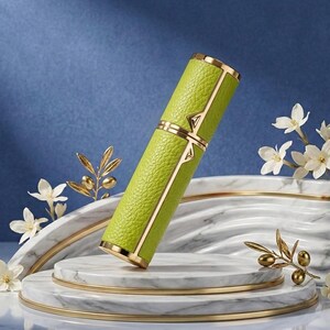 May include: A lime green, cylindrical perfume atomizer with gold accents. The atomizer has a textured leather-like exterior and is displayed on a marble and gold platform, with white flowers and gold olive branch accents.