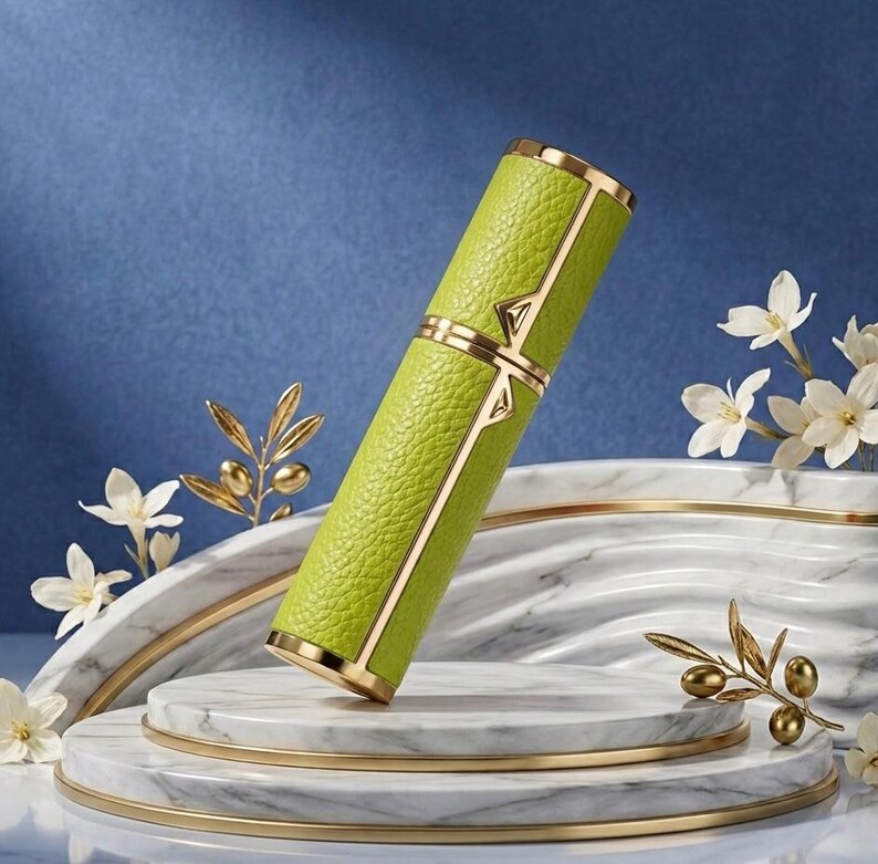 May include: A lime green, cylindrical perfume atomizer with gold accents. The atomizer has a textured leather-like exterior and is displayed on a marble and gold platform, with white flowers and gold olive branch accents.