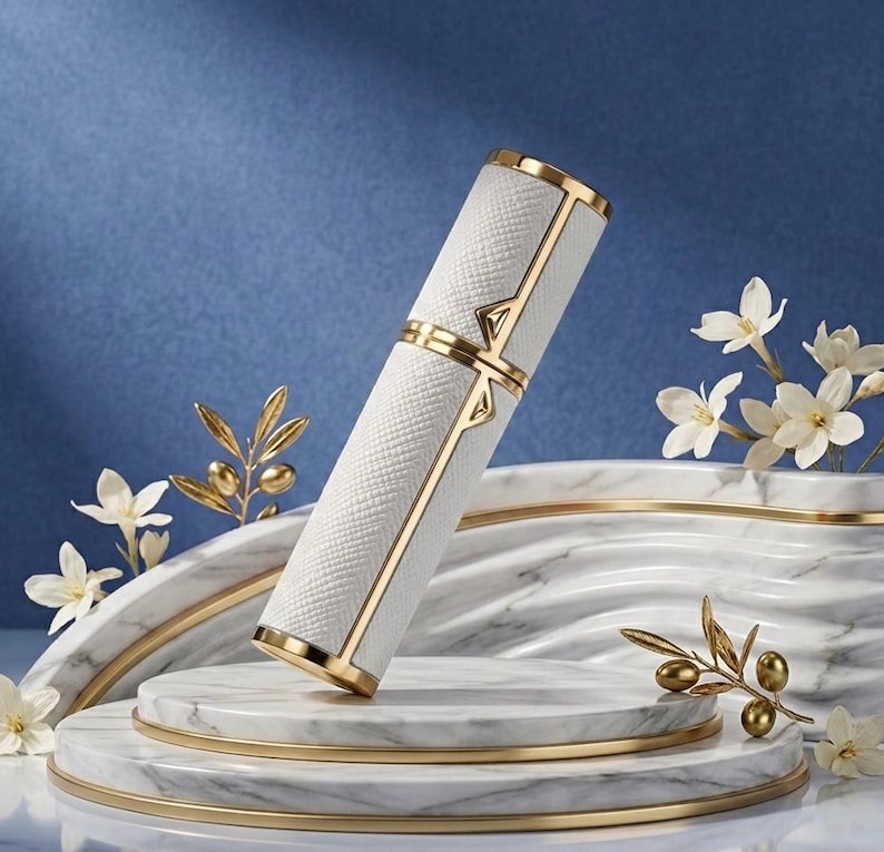 May include: A white and gold perfume atomizer with a textured surface. The cylindrical container has gold accents and a decorative design. The atomizer is displayed on a marble platform with floral accents, creating an elegant presentation.