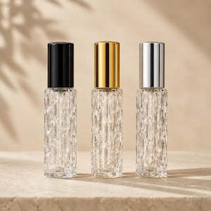 Peut inclure: Trois flacons à bille cylindriques en verre avec des surfaces texturées. Chaque flacon possède un bouchon de couleur différente : un noir mat, un doré métallique et un argenté métallique. Le verre présente un motif vertical répété.