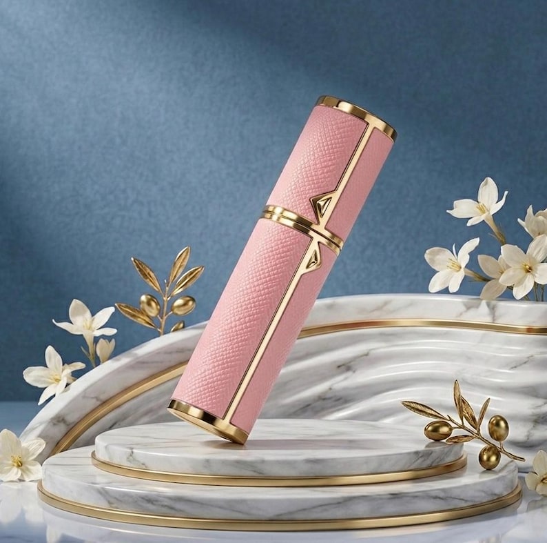 May include: A pink, rectangular perfume atomizer with gold accents. The atomizer has a textured surface and is decorated with gold trim and a gold band. The atomizer is displayed on a marble platform with white flowers and gold olive branch accents.