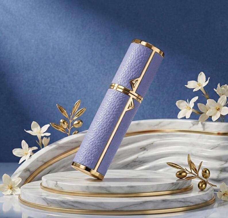 May include: A lavender-colored perfume atomizer with gold accents. The cylindrical case has a textured leather-like appearance and is trimmed with gold. The atomizer is displayed on a marble and gold tiered platform, with white flowers and gold olive branch decorations.