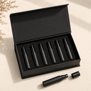 Peut inclure: Un coffret cadeau rectangulaire noir à couvercle articulé contenant six flacons pulvérisateurs cylindriques noir mat dans un insert en mousse, avec un flacon supplémentaire assorti et un bouchon noir séparé placés devant la boîte.