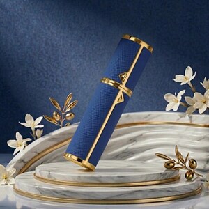 May include: A blue and gold perfume atomizer. The cylindrical case has a textured blue exterior with gold accents. The atomizer is displayed on a marble platform with floral and olive branch decorations, against a blue backdrop.