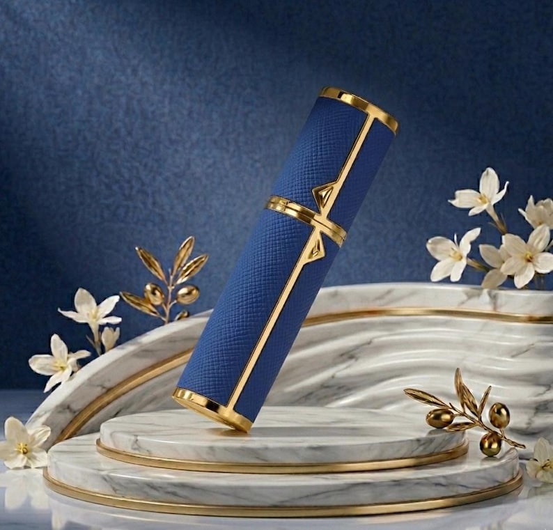 May include: A blue and gold perfume atomizer. The cylindrical case has a textured blue exterior with gold accents. The atomizer is displayed on a marble platform with floral and olive branch decorations, against a blue backdrop.
