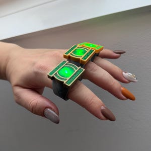 Op de afbeelding: Een ring met twee rechthoekige, goudkleurige frames met getextureerde groene randen en een centraal, cirkelvormig, oplichtend groen lichtelement. De ringband is donkergrijs en lijkt gemaakt van een mat, solide materiaal.