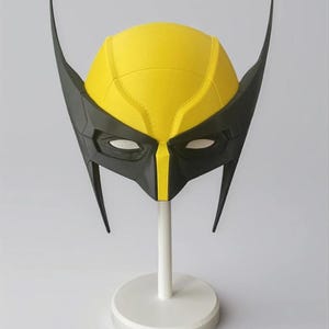 Peut inclure: Un masque de super-héros jaune et noir, présenté sur un support blanc. Le masque présente une partie supérieure jaune avec des accents noirs et des oreilles pointues. Les ouvertures des yeux sont blanches.