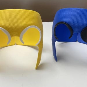 Può includere: Due maschere in stile supereroe, una gialla e una blu, con una superficie strutturata. La maschera gialla ha contorni occhi bianchi, mentre la maschera blu ha contorni occhi neri. Entrambe le maschere hanno lati lunghi e appuntiti.