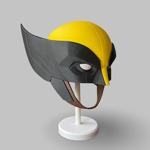 Può includere: Una maschera di Wolverine gialla e nera, con il design distintivo del personaggio. La maschera ha una finitura strutturata ed è esposta su un supporto bianco. La maschera include aperture per gli occhi e un cinturino marrone.