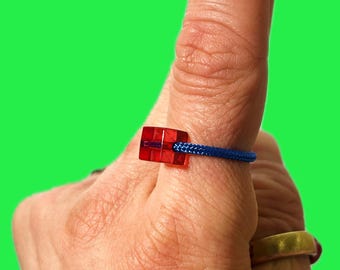 May include: A red, square-shaped bead is connected to a blue cord, forming a ring. The ring is worn on a finger against a bright green background. The ring's design is simple and modern.
